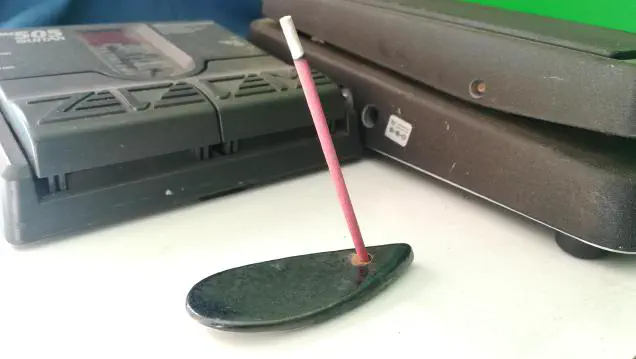 A pink stick of Nippon Kodo Rose Incense burning on a tiny droplet shaped ceramic incense holder. Surrounded by guitar effects pedals